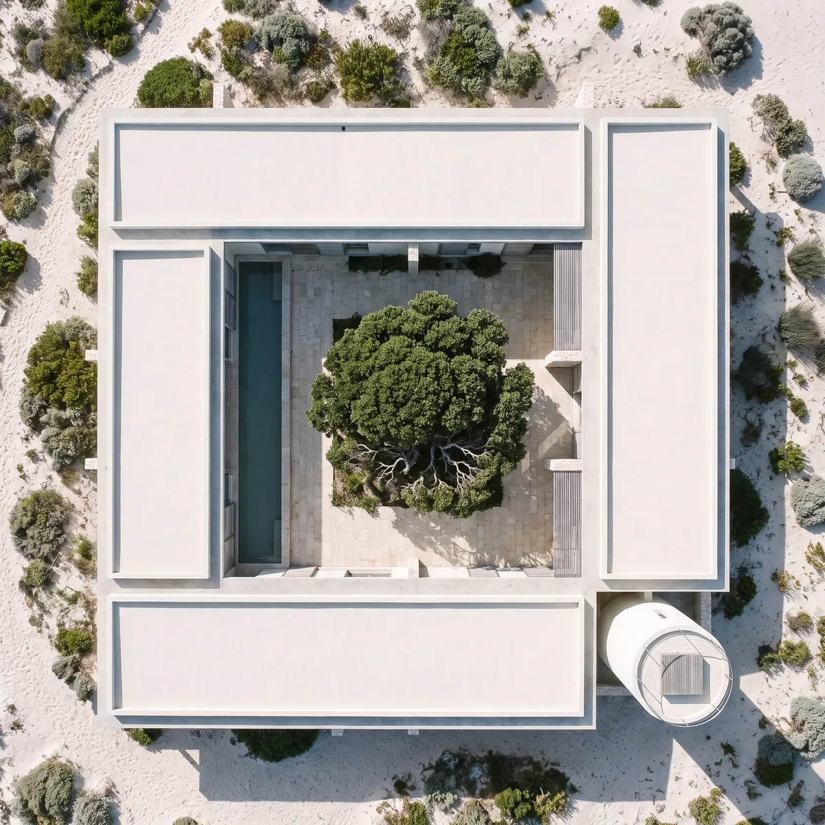 The hotel from above: four low white wings round a limestone courtyard with an old milkwood tree and a long pool, the round tower at one corner, in the dunes